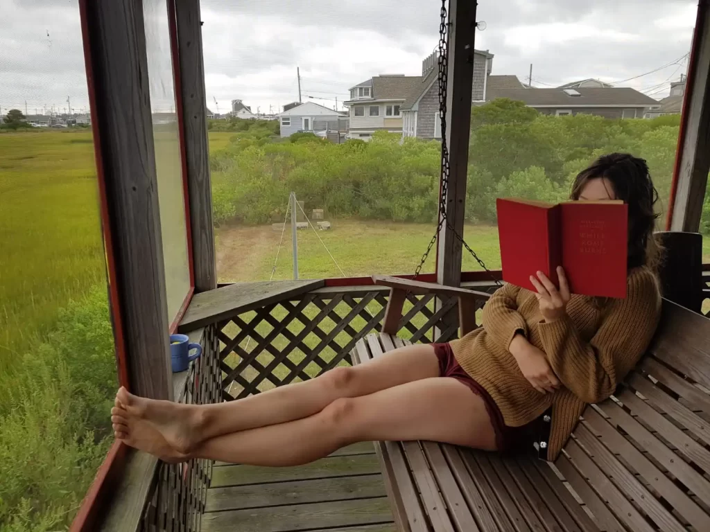 Woman sitting with legs stretched out on bench reading a book with a view of grass out the window